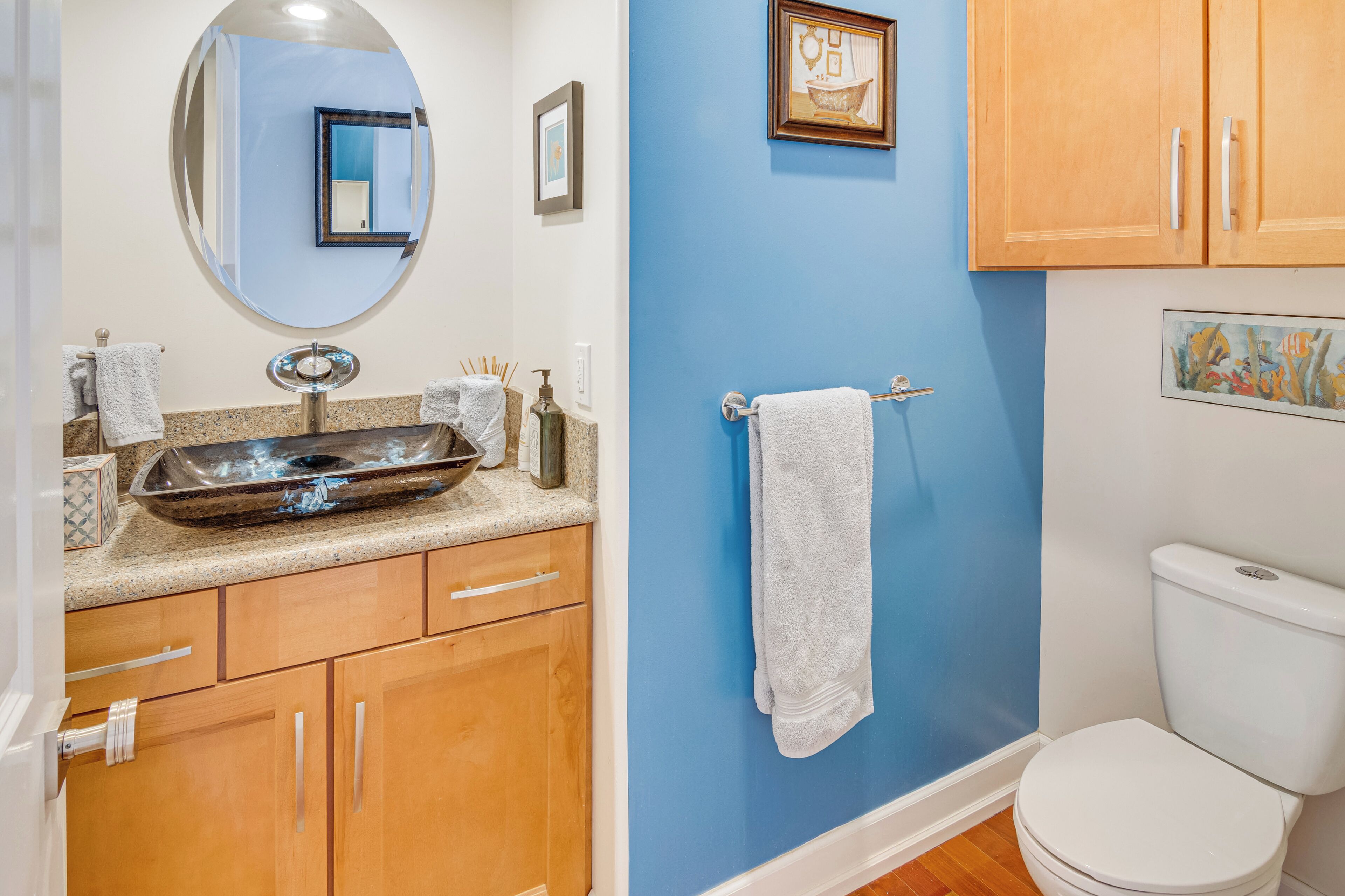 Un bagno con parete blu, mobili in legno, un water bianco e uno specchio con un quadro incorniciato sopra.