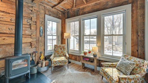 Interior rústico de una cabaña con estufa de leña, dos sillones con estampado floral, una mesita con lámpara y amplias ventanas que ofrecen vistas al exterior.