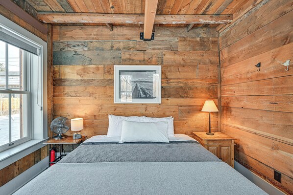 Camera in legno con un letto, due lampade da comodino, una ventola, una foto incorniciata appesa al muro e una finestra.