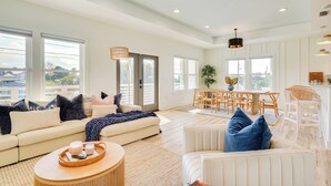 Een ruime woonkamer met een bank, fauteuils en een salontafel. Er is een eethoek met een tafel en stoelen, en een keuken met een aanrecht en barkrukken.
