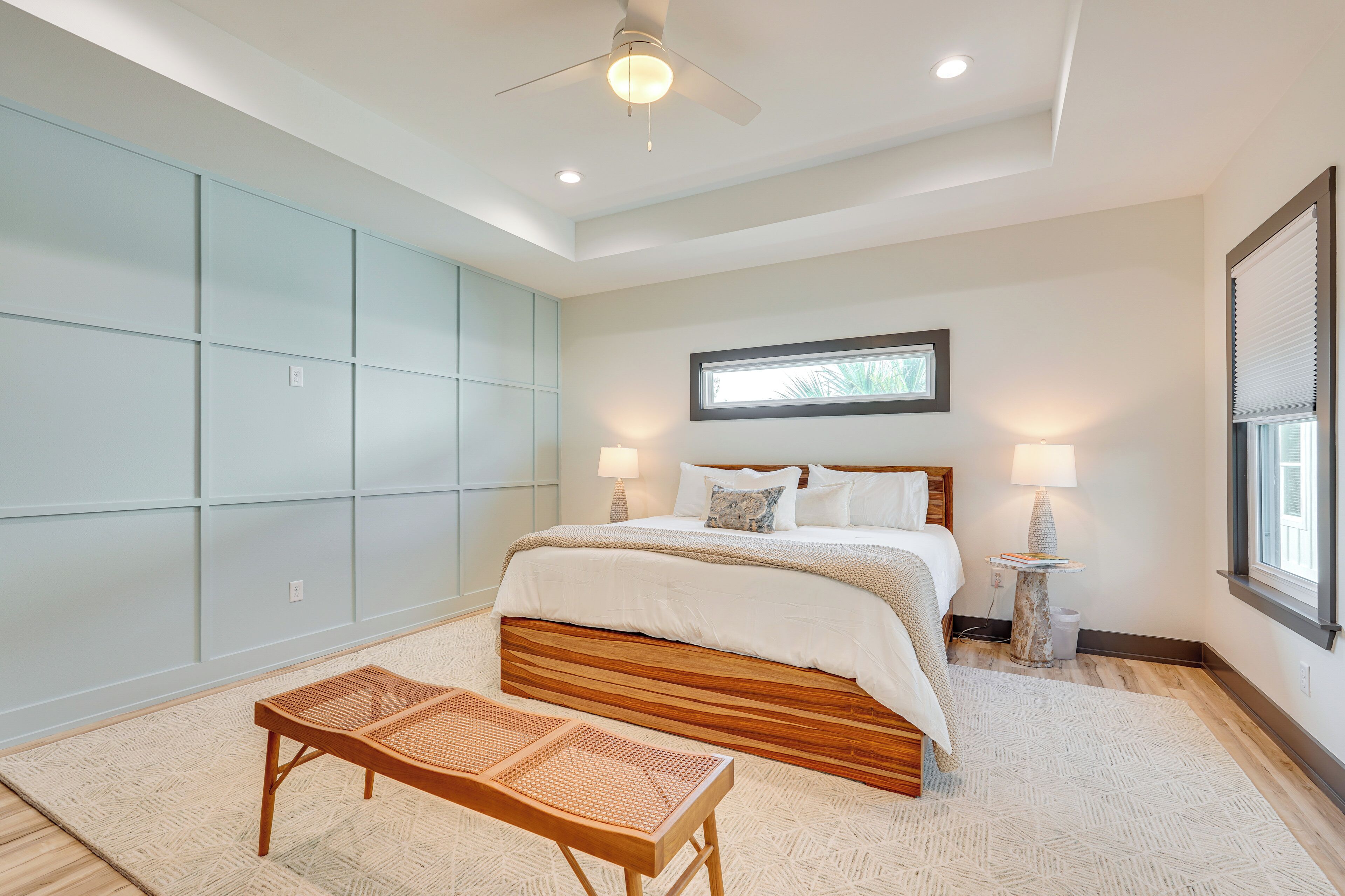 A bedroom with a large bed, a ceiling fan, a window with blinds, and a bedside table with a lamp.