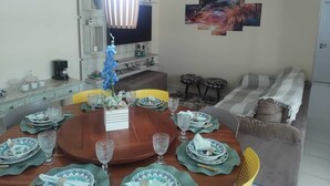 A dining area with a round wooden table set for four, surrounded by chairs. A television is mounted on the wall above the table, and there is a sofa with a patterned throw blanket in the adjacent living area. - Ground floor apartment, in the VG Sun condominium. Condominium right on the sand. (Caucaia)