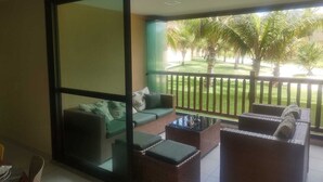 A balcony with wicker furniture, a glass partition, and a view of a well-maintained lawn and palm trees. - Ground floor apartment, in the VG Sun condominium. Condominium right on the sand. (Caucaia)
