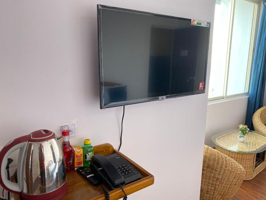 A room with a flat-screen TV mounted on the wall, a wooden tray with a kettle, condiments, and a telephone. There is a wicker chair and a small table with a plant.