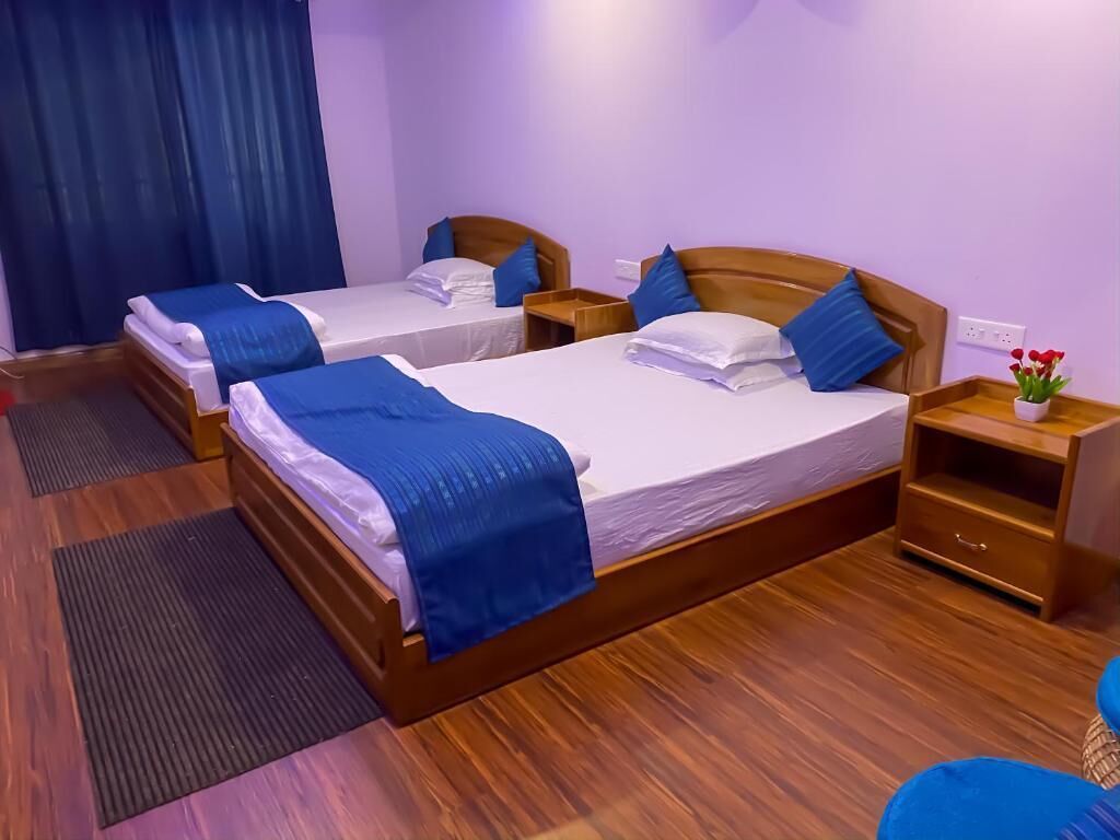 Quarto de hotel com duas camas de madeira, cada uma com roupa de cama e almofadas azuis, uma mesa de cabeceira de madeira, uma pequena mesa com uma planta e piso de madeira.
