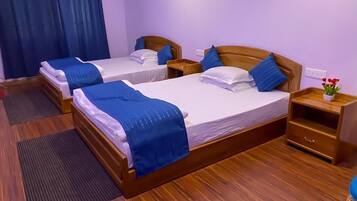 Quarto de hotel com duas camas de madeira, cada uma com roupa de cama e almofadas azuis, uma mesa de cabeceira de madeira, uma pequena mesa com uma planta e piso de madeira.