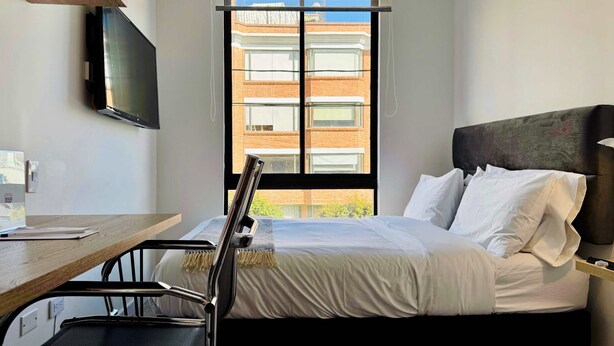 A hotel room with a bed, a desk, a chair, a television, and a window with a view of a brick building. - w Radiant 1BR in Santa Barbara (Bogotá)