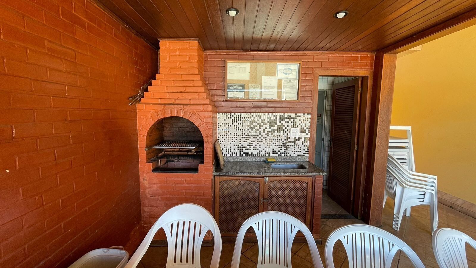 Un horno de ladrillo con salpicadero de azulejos, un lavamanos y una pequeña encimera. Se ven sillas de plástico blanco y una puerta de madera al fondo.