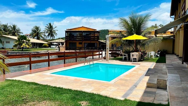 A swimming pool with a wooden deck and a stone border, surrounded by palm trees and a building with a tiled roof.