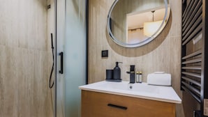 Ein modernes Badezimmer mit Duschkabine aus Glas, einem runden Spiegel, einem weißen Waschbecken mit Holzschrank und einem Handtuchhalter.