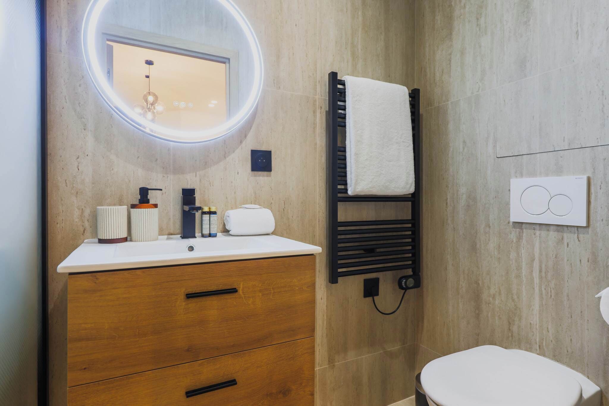 Un baño moderno con un mueble de tocador de madera, un espejo redondo con luz, un perchero y un inodoro.