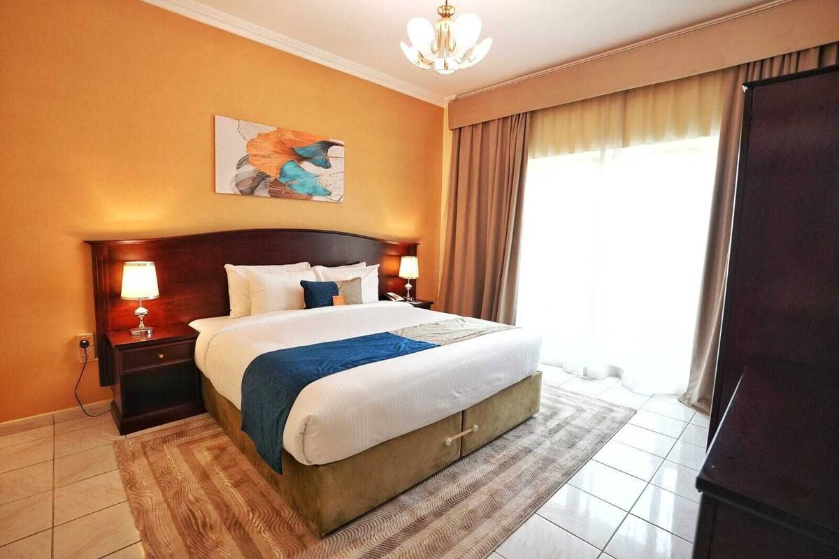 A hotel room with a large bed, two bedside tables with lamps, a painting on the wall, and a view of the outdoors through the window.