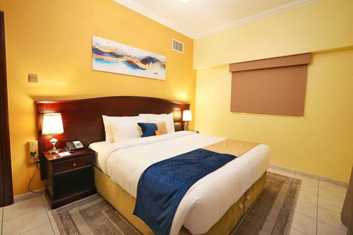 A hotel room with a large bed, two bedside tables with lamps, a painting on the wall, and a window with a blind.