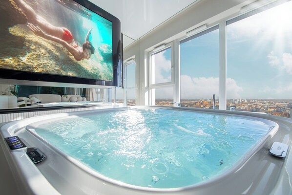 A modern indoor swimming pool with a large TV screen displaying an underwater scene, large windows offering a city view, and a control panel for pool features.
