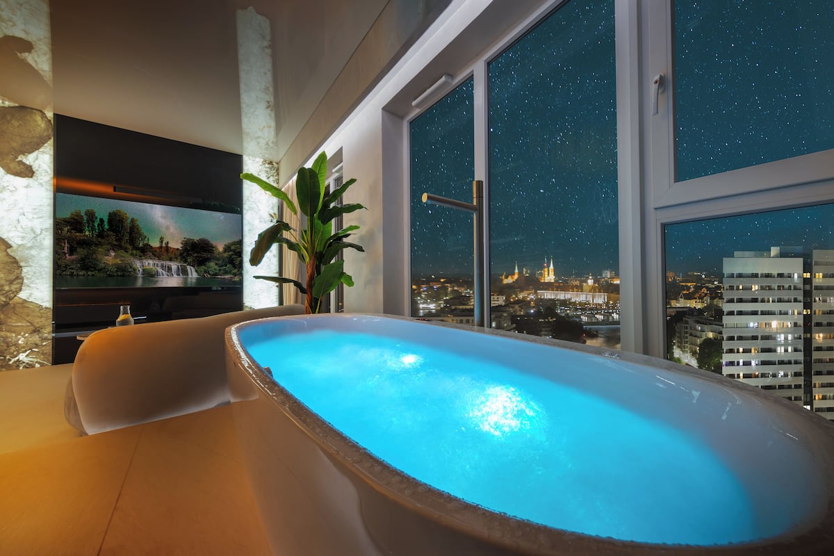 Ein modernes Badezimmer mit einer großen Badewanne, einem Blumentopf und Blick auf die Stadt durch bodengrosse Fenster.