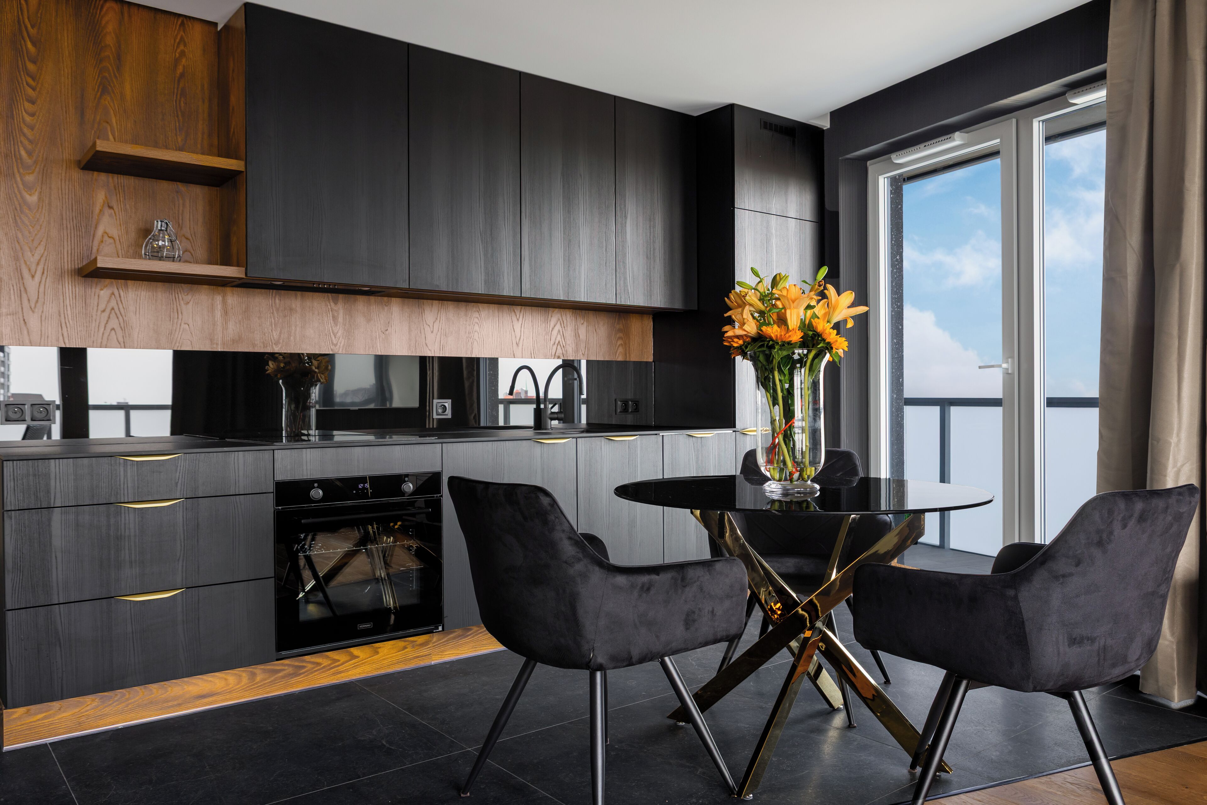 A modern kitchen with dark cabinetry, a black oven, and a dining area with two black chairs and a glass table.