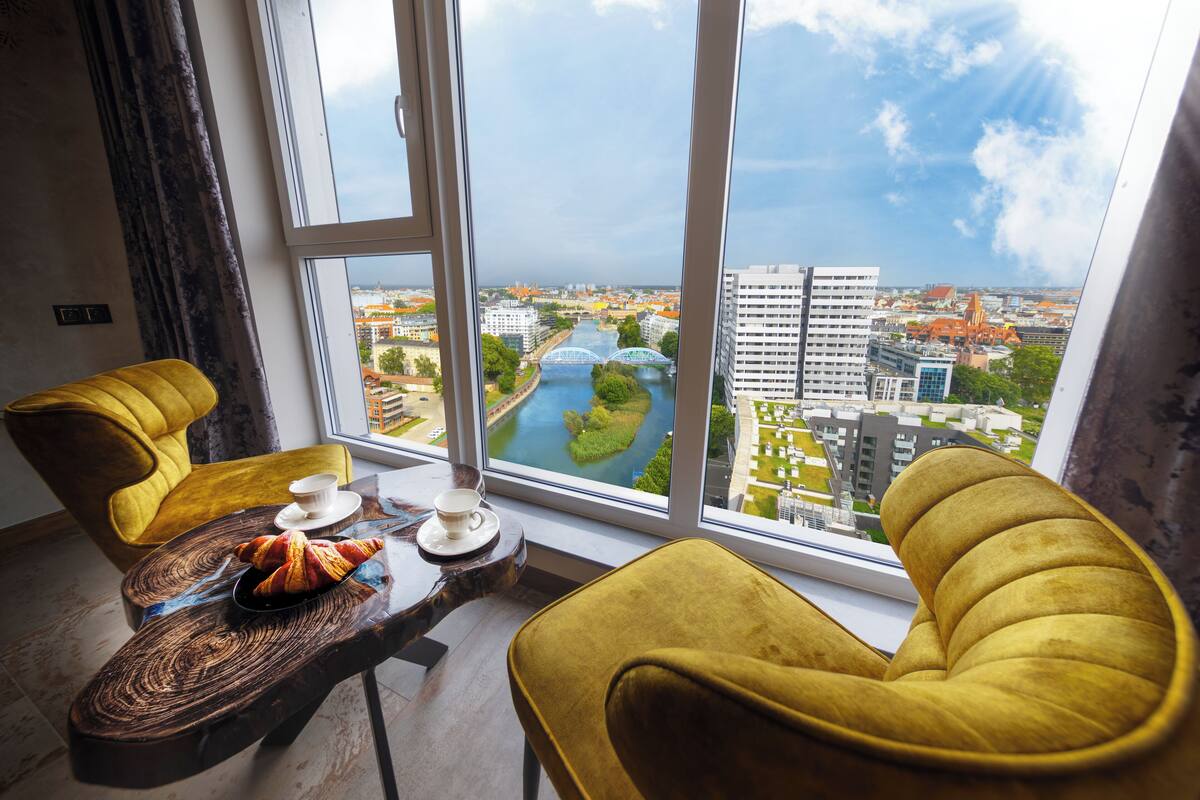 Ein modernes Hotelzimmer mit einem großen Fenster, das einen Blick auf eine Stadtlandschaft und einen Fluss bietet. Zwei gelbe Sessel und ein Holztisch mit Croissants und einer Tasse sind vorhanden.