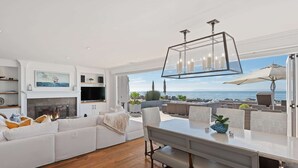 Un salon moderne avec un grand canapé d’angle blanc, un coin repas avec un banc et une vue sur l’océan.