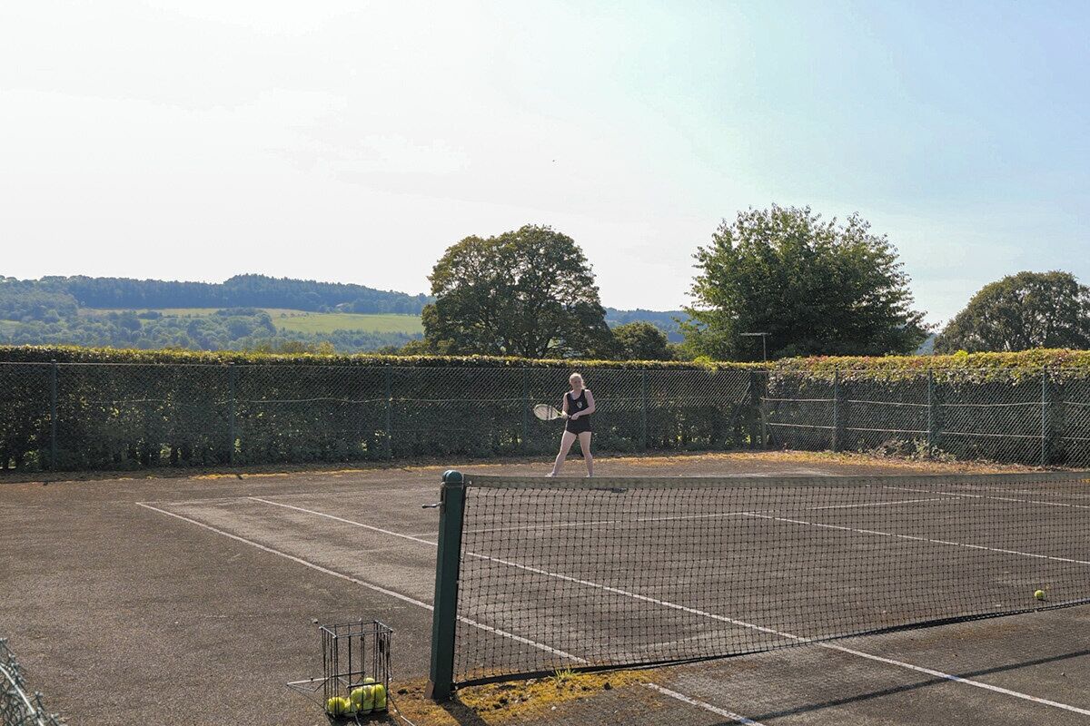 Ein Tennisplatz mit Netz, ein Spieler in Schwarz und ein Ballständer mit Tennisbällen.