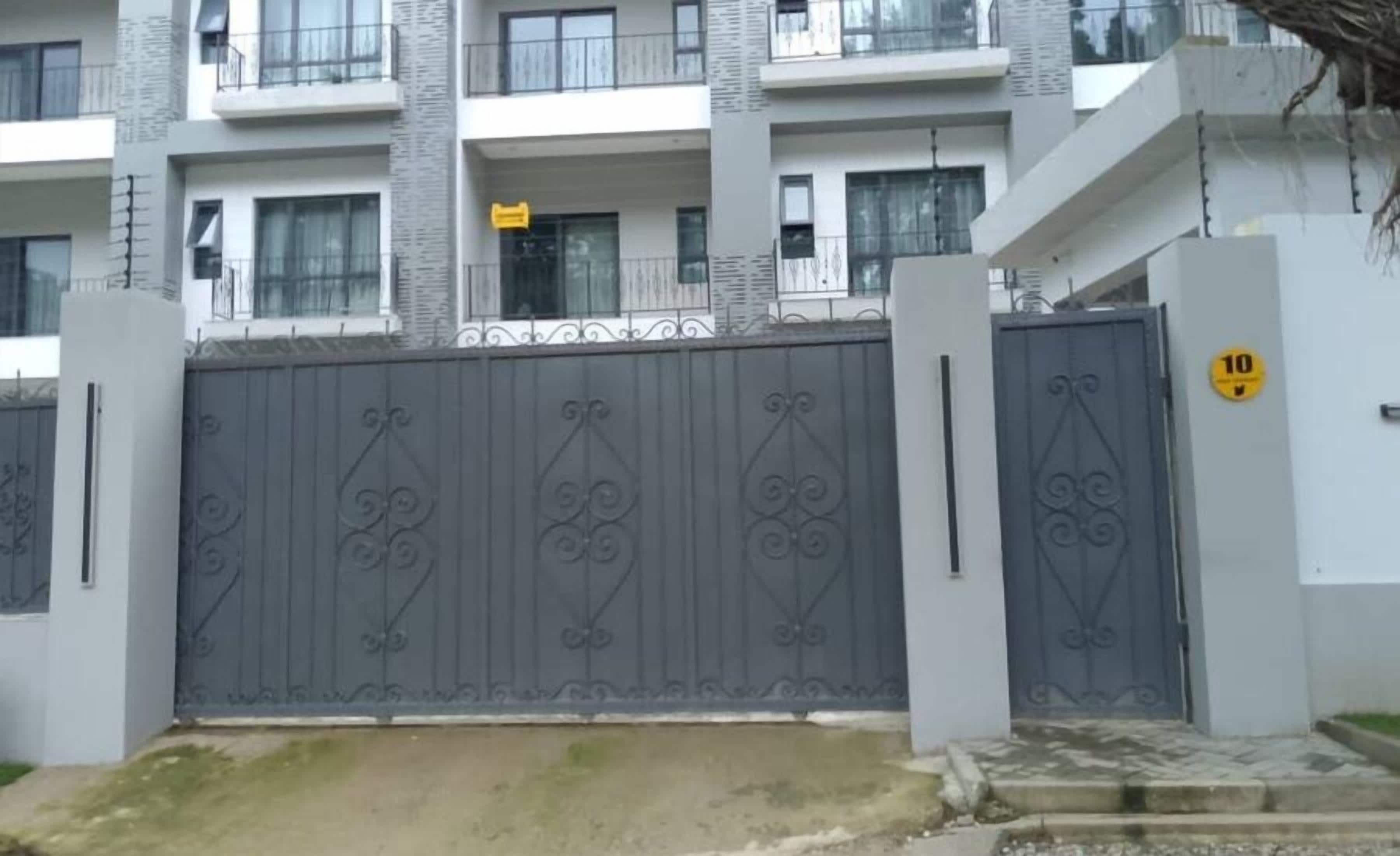 Une entrée sécurisée, avec portail, donne accès à un immeuble résidentiel à plusieurs étages, doté d’une architecture moderne, de balcons et d’un portail imposant agrémenté d’éléments décoratifs.