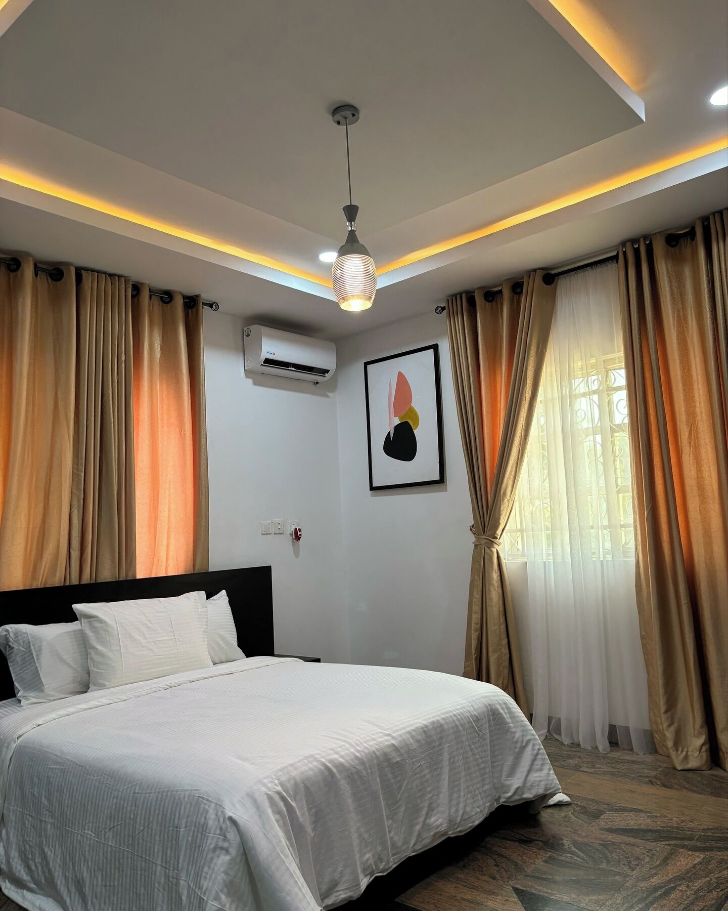 A bedroom with a bed, curtains, an air conditioning unit, a ceiling light, and a wall-mounted artwork.