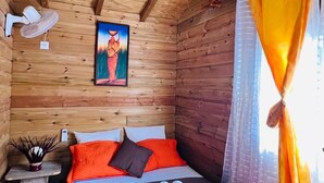 Habitación de cabaña de madera con una cama, una mesita de noche, un ventilador fijado a la pared, un cuadro y una ventana con cortinas amarillas.