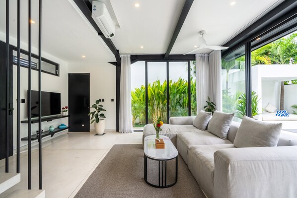 A modern living room with a large sofa, a coffee table, and a TV on a black stand. There are potted plants and a view of greenery outside.
