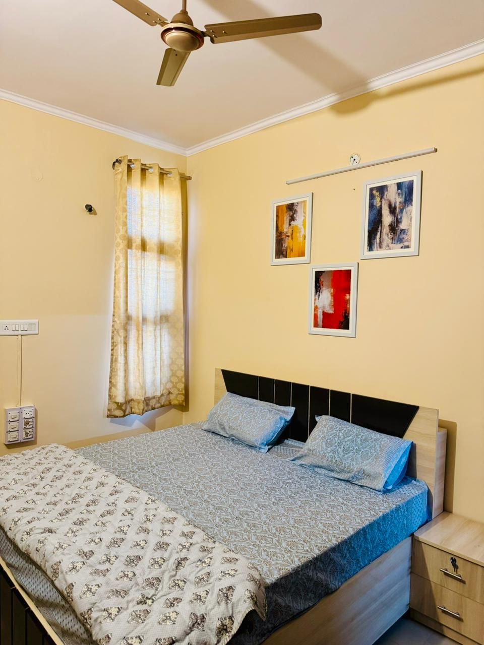 Un dormitorio con una cama, ventilador de techo, mesita de noche y una pared con tres cuadros.