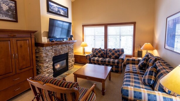 A cozy living room with a stone fireplace, plaid sofas, a wooden coffee table, and a television mounted above the fireplace. - 3323 - Two Bedroom Standard Powderhorn Lodge 2 Condo (Salt Lake City)