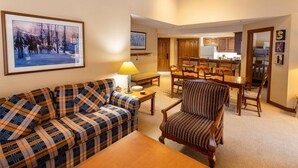 A cozy living room with a plaid sofa, a patterned armchair, and a wooden coffee table. There is a dining area with wooden chairs and a table, and a kitchen in the background. - 3323 - Two Bedroom Standard Powderhorn Lodge 2 Condo (Salt Lake City)