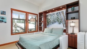 A bedroom with a bed, a nightstand, a lamp, a window with a view of a snowy landscape, and a shelf with decorative items. - 1311 - Three Bedroom Deluxe Eagle Springs West 3 Condo (Solitude)