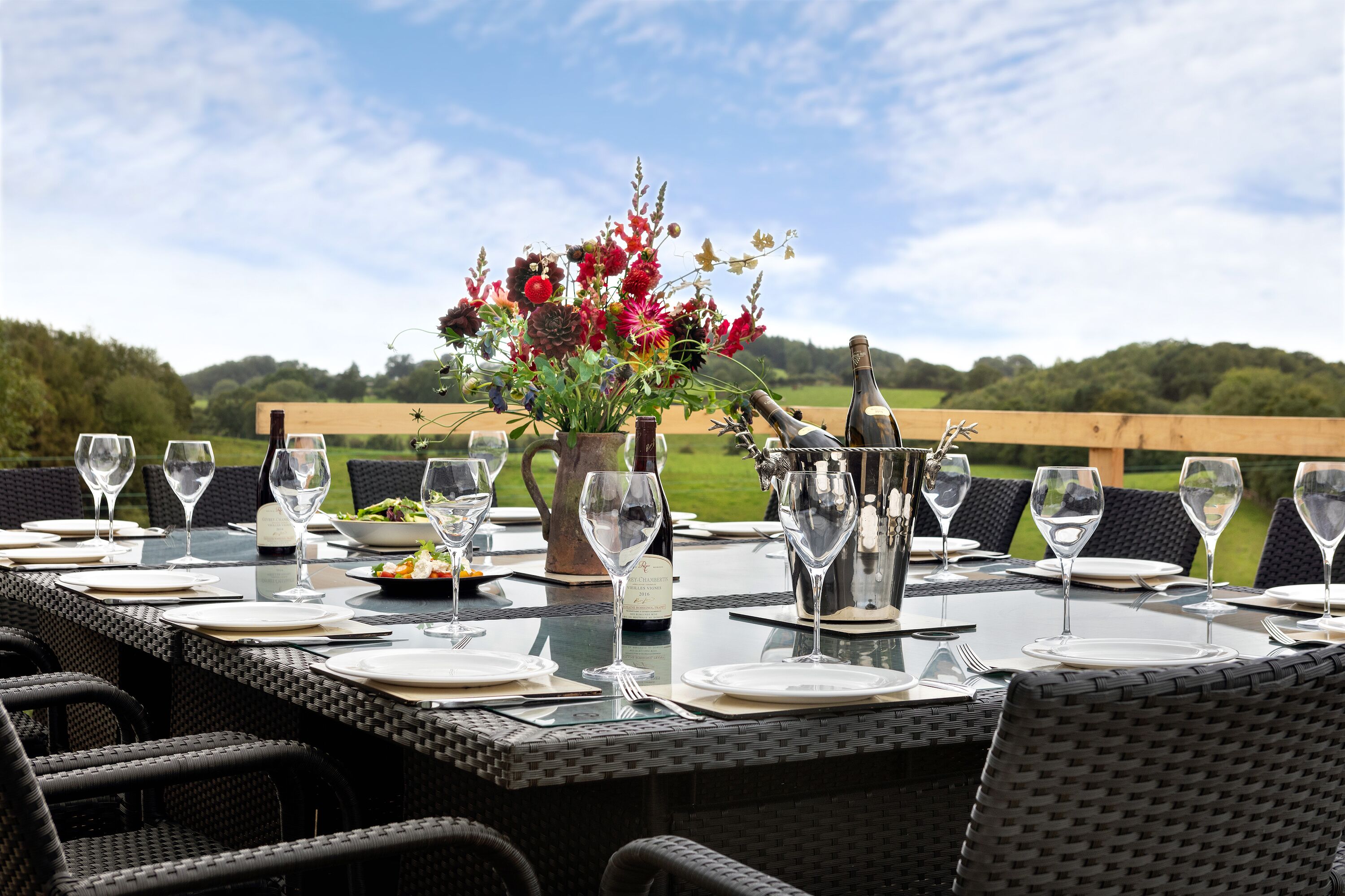 Una mesa para comer al aire libre con platos blancos, copas de vino y un centro de mesa floral.
