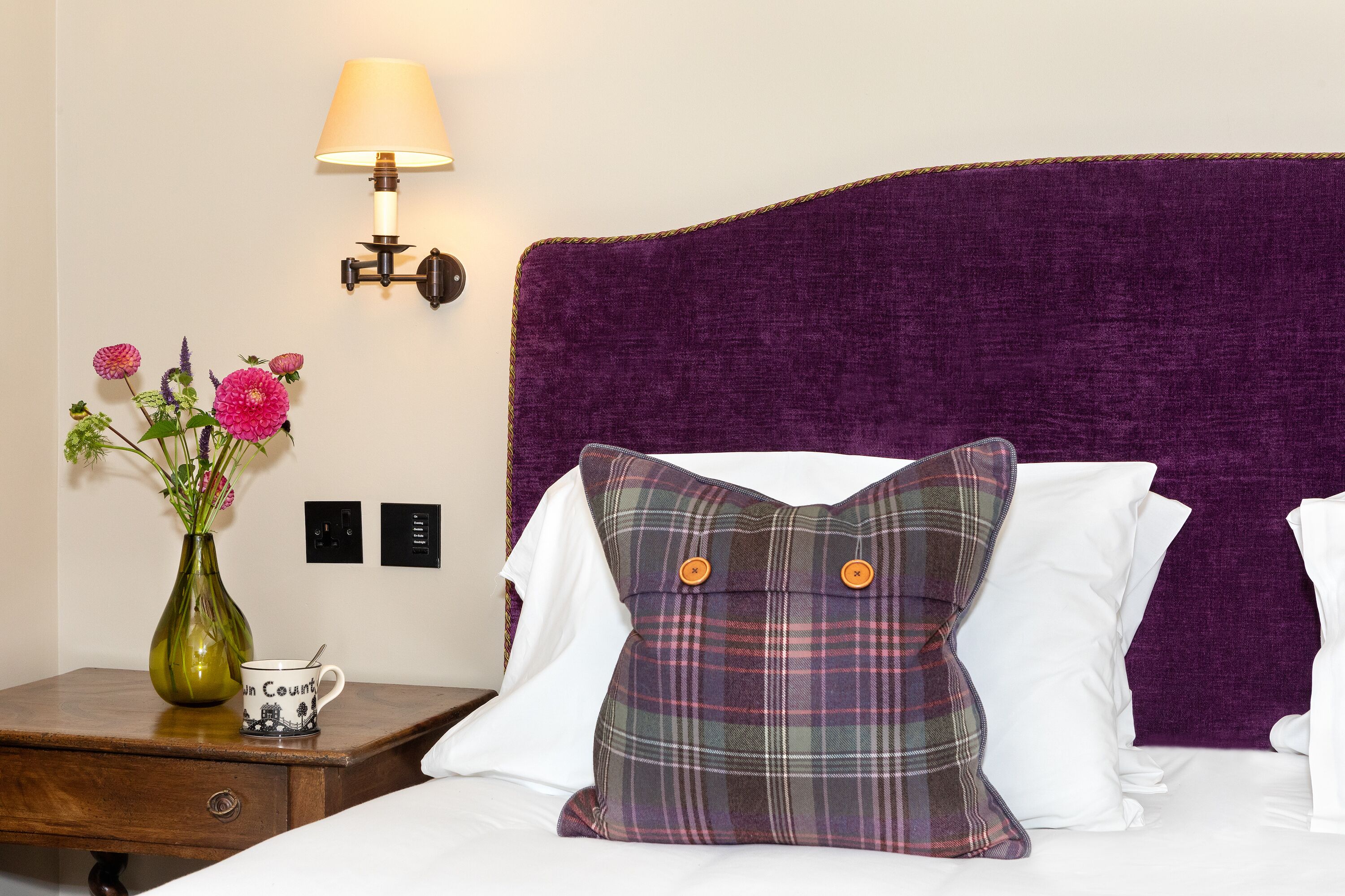 Una cama bien hecha con un cabecero morado, una almohada escocesa, un florero con flores y una mesita de noche con una lámpara y una taza.