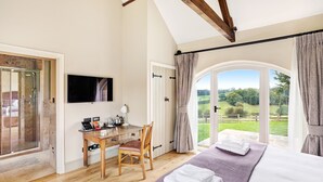 Un dormitorio con una cama grande, un escritorio con silla, un televisor y vistas a un paisaje verde a través de una puerta de cristal.