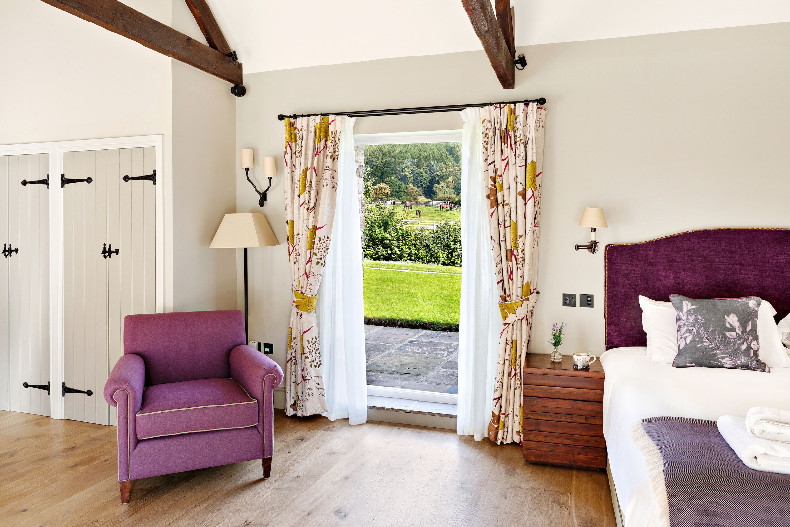 Una habitación con cama, una silla, un ventanal con cortinas y vistas a un jardín.