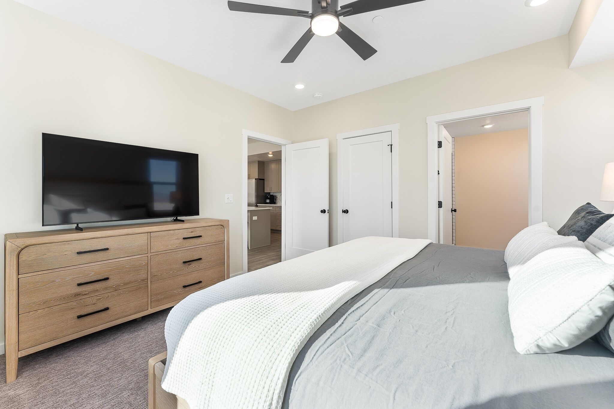 A bedroom with a bed, a TV on a wooden dresser, a ceiling fan, and a door leading to a bathroom.