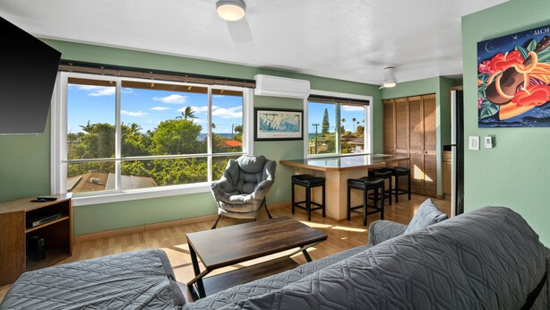 A room with a bed, a desk, a chair, and large windows offering a view of greenery and buildings. - Kauai Hale Kahanalu 8 2 Bedroom Condo (Koloa)