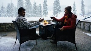 Dos personas sentadas en una mesa al aire libre con una fogata, árboles nevados al fondo.