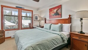 Un dormitorio con una cama grande, un ventilador de techo, una mesita de noche, una lámpara, un cuadro y una ventana con vistas a árboles.