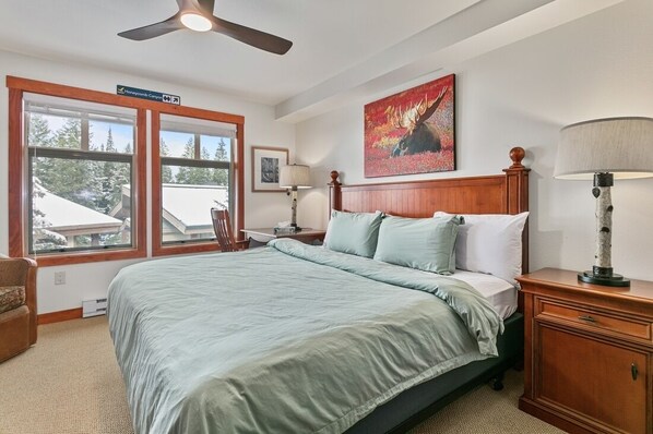 A bedroom with a large bed, a ceiling fan, a nightstand, a lamp, a painting, and a window with a view of trees. - 1311b - King Standard Eagle Springs West Hotel Room (Solitude)