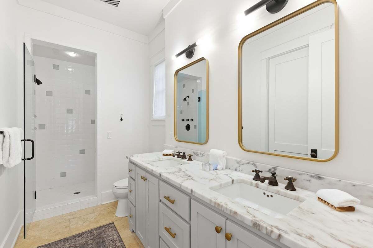 Een moderne badkamer met een douchecabine van glas, een wit toilet, een marmeren wastafel met gouden details en twee grote spiegels met gouden lijstwerk.