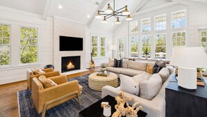 A spacious living room with a fireplace, large windows, and a modern chandelier. - Watersound Beach! With Carriage House, Private Pool, LSV/Golf Cart, & 4 Bikes! (Watersound)