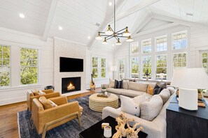 A spacious living room with a fireplace, large windows, and a modern chandelier.