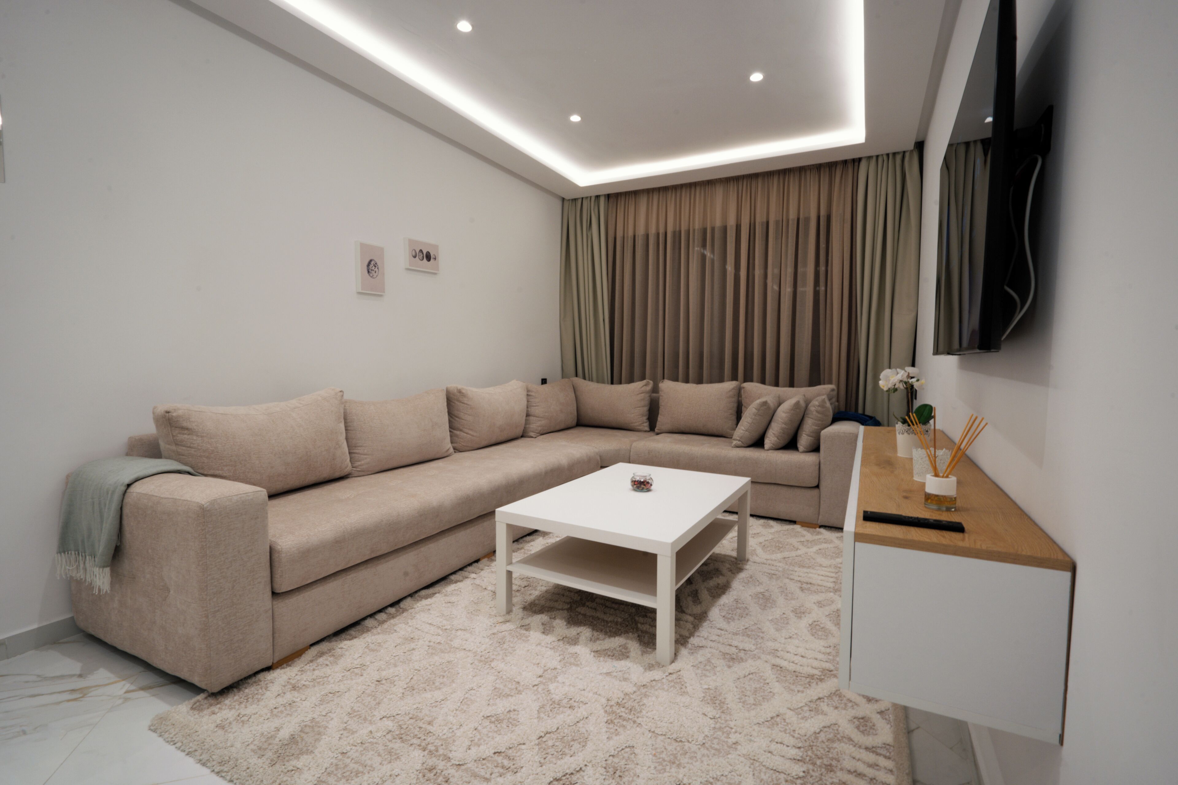 Un salon moderne avec un canapé beige, une table basse blanche et une table d’appoint en bois, sur le mur étant fixé un téléviseur.