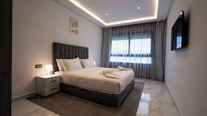 Een moderne hotelkamer met een groot bed, een nachtkastje met een lamp, een flatscreen televisie aan de muur en een groot raam met gordijnen.