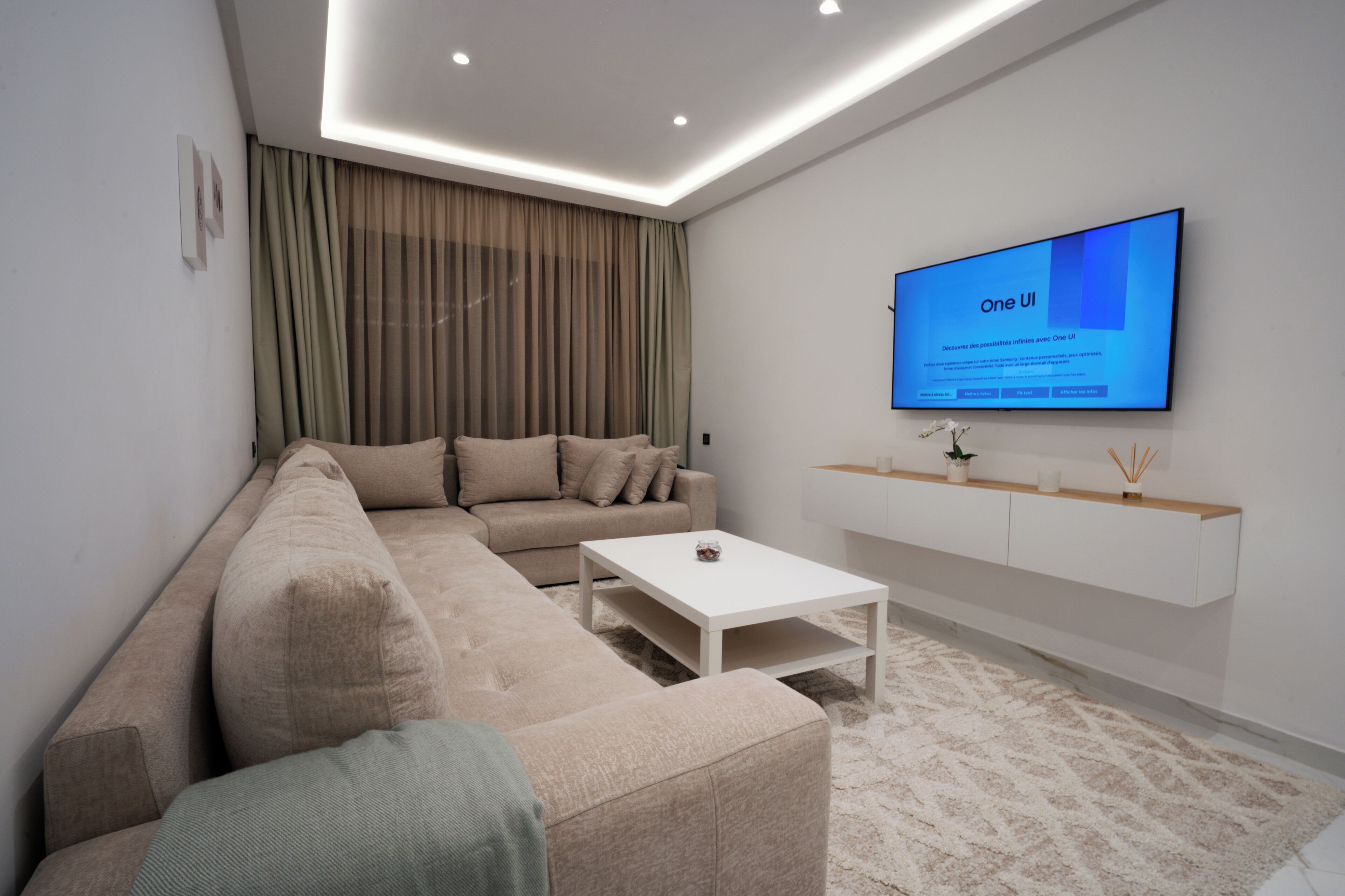 Een moderne woonkamer met een beige modulaire bank, een witte salontafel en een tv met daarop 'One UI'.