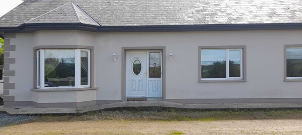 Une maison à deux étages avec une porte blanche, deux fenêtres au rez-de-chaussée et un toit gris.