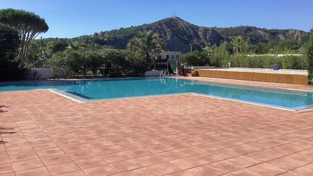 A swimming pool with tiled flooring, surrounded by greenery and a hill in the background.