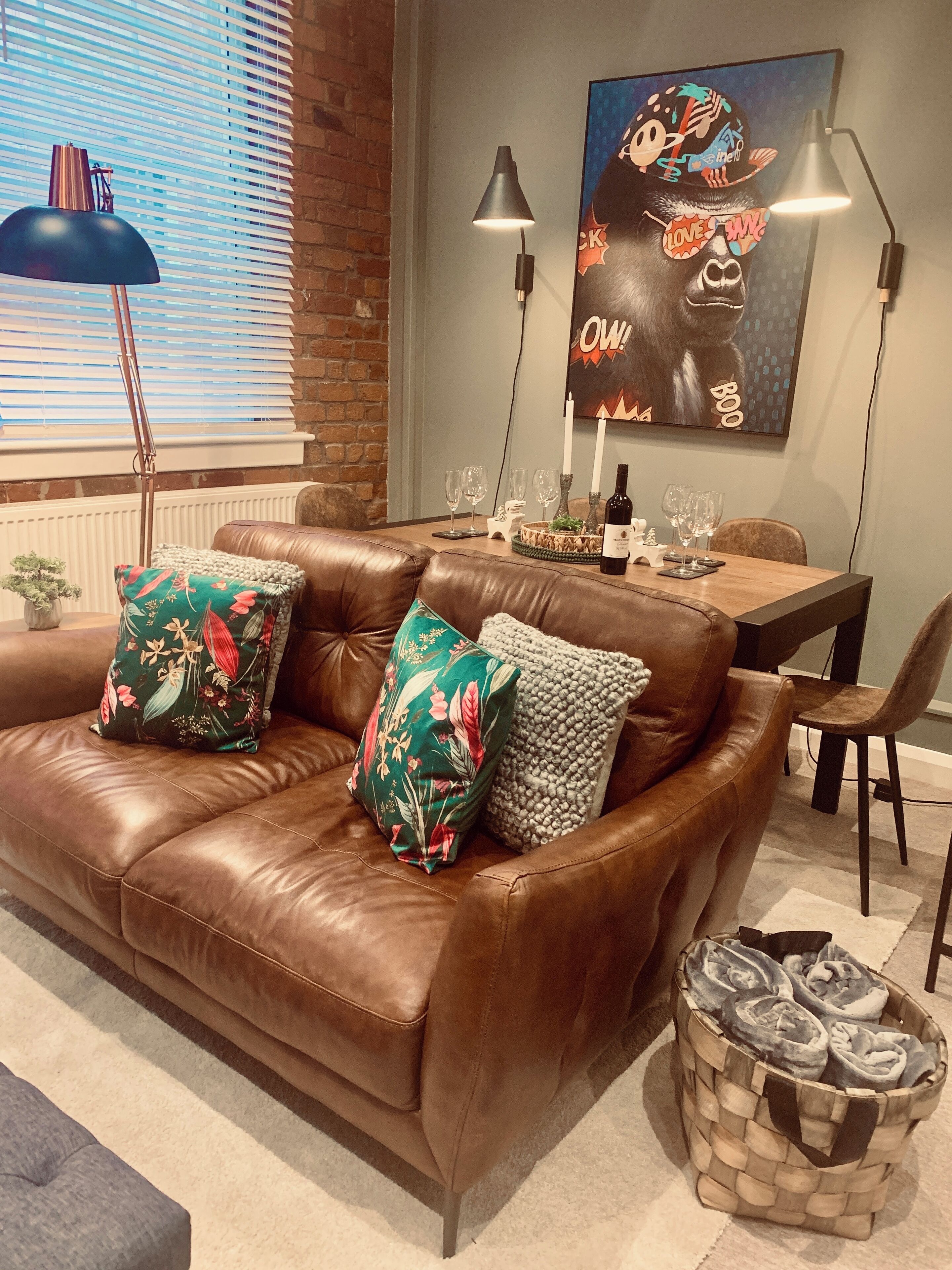A brown leather sofa with floral and textured pillows, a dining table with wine glasses and a bottle, and a wall with a colorful poster.