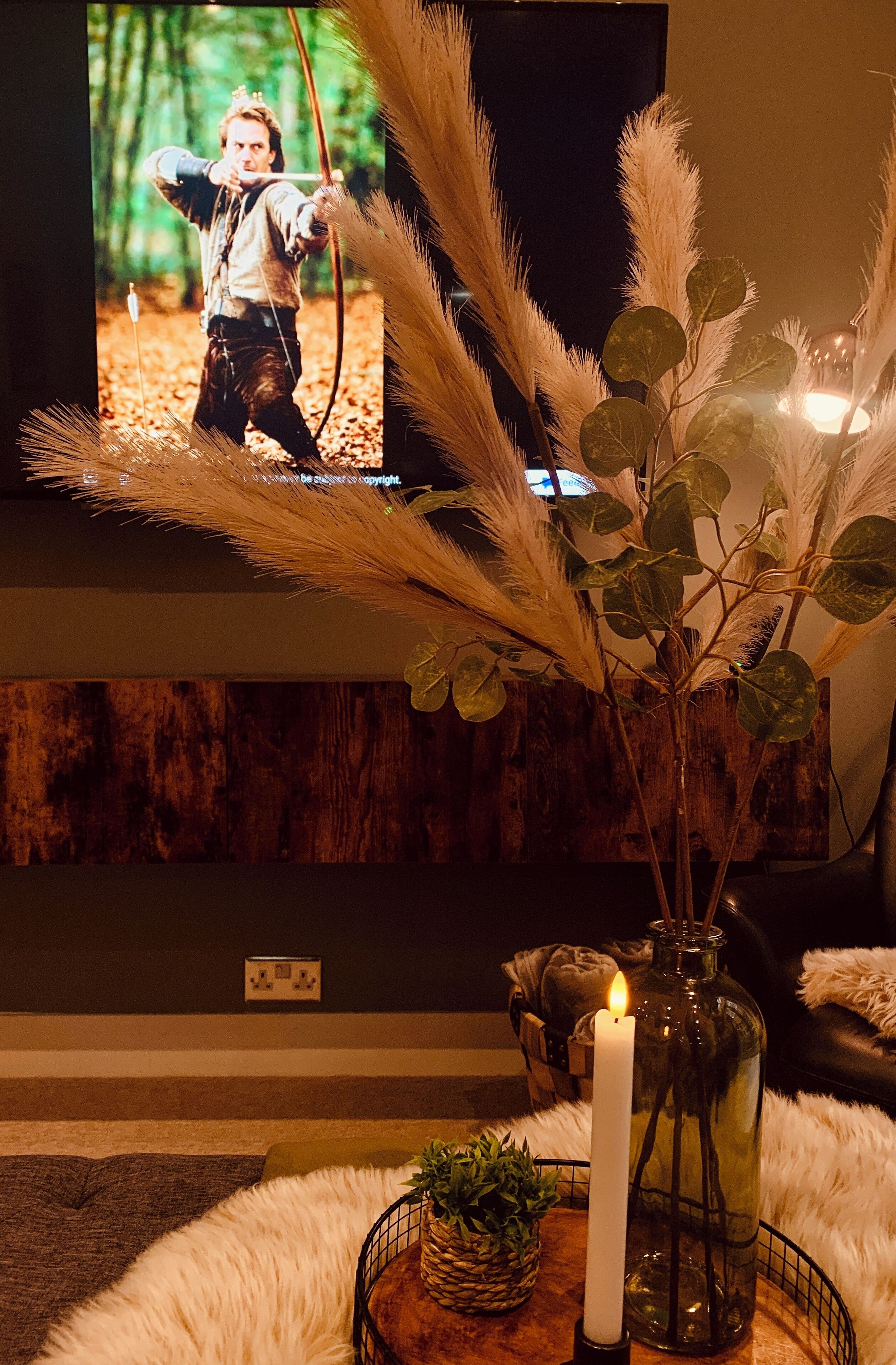 A cozy living room with a television displaying a movie scene, a lit candle, and decorative plants.